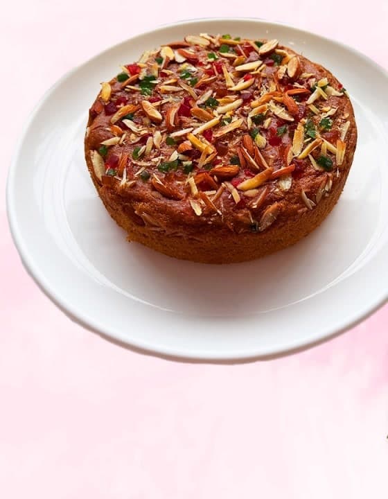 Eggless mango cake topped with almonds, tutti frutti and dry fruits on a white plate